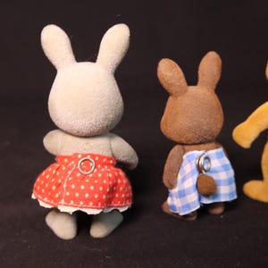 Może przedstawiać: Trzy figurki zabawek kr&oacute;lik&oacute;w. Jeden jest jasnoszary z czerwoną sp&oacute;dnicą w kropki. Drugi jest brązowy z niebiesko-białymi szortami w kratkę. Trzeci jest ż&oacute;łty. Wszystkie mają puszystą fakturę.