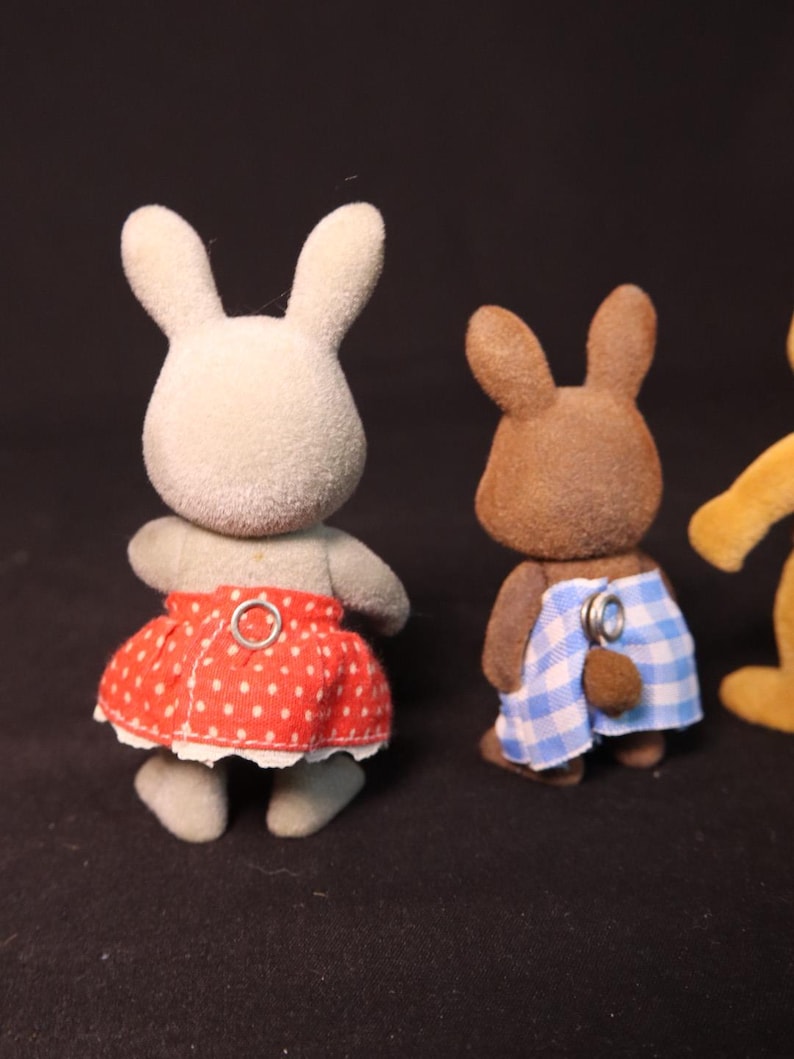 Może przedstawiać: Trzy figurki zabawek kr&oacute;lik&oacute;w. Jeden jest jasnoszary z czerwoną sp&oacute;dnicą w kropki. Drugi jest brązowy z niebiesko-białymi szortami w kratkę. Trzeci jest ż&oacute;łty. Wszystkie mają puszystą fakturę.