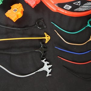 May include: A collection of colorful toy accessories, including ripcords and launchers. The ripcords are in various colors: black, yellow, green, tan, blue, and red. The launchers have different designs and colors, with an orange piece visible. The items are on a black surface.