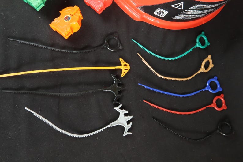 May include: A collection of colorful toy accessories, including ripcords and launchers. The ripcords are in various colors: black, yellow, green, tan, blue, and red. The launchers have different designs and colors, with an orange piece visible. The items are on a black surface.