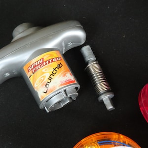 May include: A gray Spin Fighter launcher toy with a spring and two colorful spinning tops. The launcher has a handle and a cylindrical base with the words "Spin Fighter Launcher" printed on it.