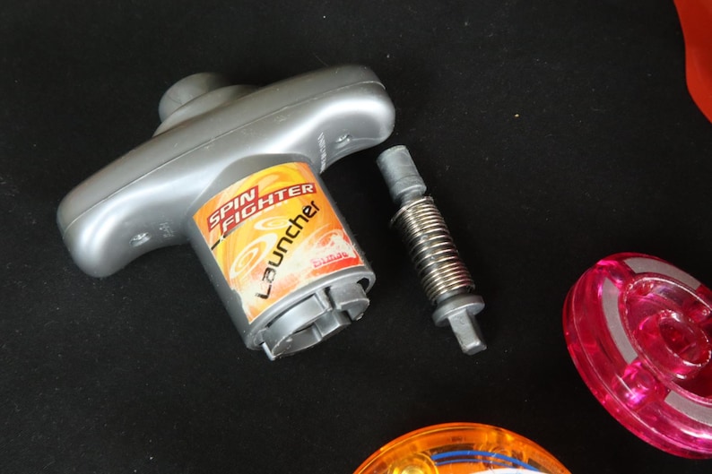 May include: A gray Spin Fighter launcher toy with a spring and two colorful spinning tops. The launcher has a handle and a cylindrical base with the words "Spin Fighter Launcher" printed on it.