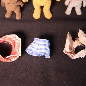 Może przedstawiać: Kolekcja miniaturowych pluszowych misi&oacute;w i ubranek dla lalek. Ubranka obejmują czerwoną i białą czapkę w kropki, niebiesko-białe szorty w kratkę oraz r&oacute;żowo-biały szalik z falbankami. Misie są w r&oacute;żnych kolorach.