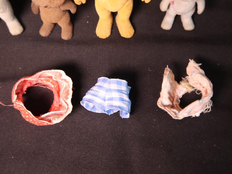 Może przedstawiać: Kolekcja miniaturowych pluszowych misi&oacute;w i ubranek dla lalek. Ubranka obejmują czerwoną i białą czapkę w kropki, niebiesko-białe szorty w kratkę oraz r&oacute;żowo-biały szalik z falbankami. Misie są w r&oacute;żnych kolorach.