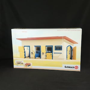 Può includere: Un set da gioco con scuderia per cavalli Schleich in una scatola rettangolare. La scatola mostra un edificio beige con tetto rosso, finiture blu e porte gialle. Il testo 40164 e Schleich S sono visibili. Le dimensioni sono 54,5 cm x 21 cm x 22 cm.