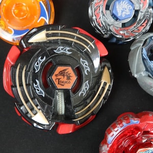 May include: A collection of colorful spinning tops, including a black and red top with the word "Draco" and a dragon emblem. Other tops feature blue, orange, and silver designs. These toys are designed for competitive play.