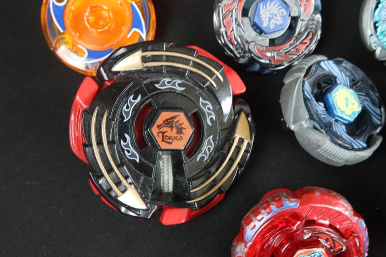 May include: A collection of colorful spinning tops, including a black and red top with the word "Draco" and a dragon emblem. Other tops feature blue, orange, and silver designs. These toys are designed for competitive play.