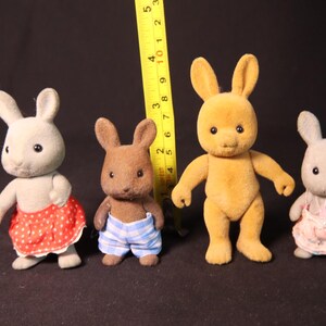 Może przedstawiać: Cztery figurki kr&oacute;lik&oacute;w-zabawek w r&oacute;żnych kolorach, w tym szarym, brązowym i ż&oacute;łtym. Kr&oacute;liki mają na sobie stroje, takie jak sp&oacute;dnice i szorty. Za figurkami umieszczono ż&oacute;łtą miarkę.