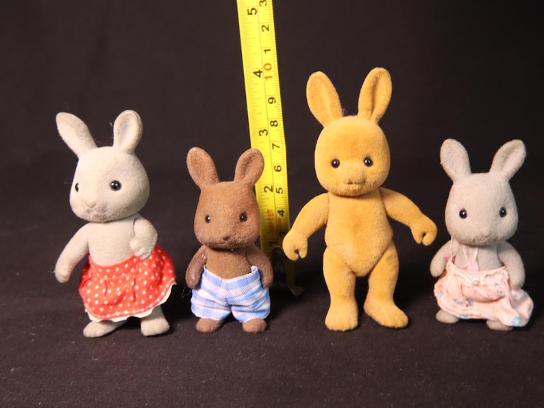 Może przedstawiać: Cztery figurki kr&oacute;lik&oacute;w-zabawek w r&oacute;żnych kolorach, w tym szarym, brązowym i ż&oacute;łtym. Kr&oacute;liki mają na sobie stroje, takie jak sp&oacute;dnice i szorty. Za figurkami umieszczono ż&oacute;łtą miarkę.