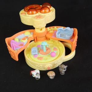 May include: A yellow and orange toy playset with a carousel design, featuring a central platform, three side compartments, and three small animal figurines. The playset has a flower and leaf design.