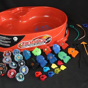 May include: A red plastic Beyblade XTS Extreme Top System arena with various colorful tops, launchers, and accessories. The set includes tops in blue, green, orange, and red, along with ripcords and other components. The packaging indicates the toy is for ages 8+.