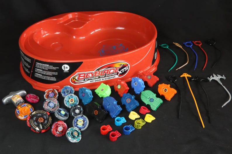 May include: A red plastic Beyblade XTS Extreme Top System arena with various colorful tops, launchers, and accessories. The set includes tops in blue, green, orange, and red, along with ripcords and other components. The packaging indicates the toy is for ages 8+.