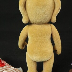 May include: A tan, flocked, anthropomorphic animal figurine standing upright. The figure has rounded ears, a smooth, textured surface, and a simple, upright posture. Beside the figurine lies a small, striped fabric garment with a red bow.