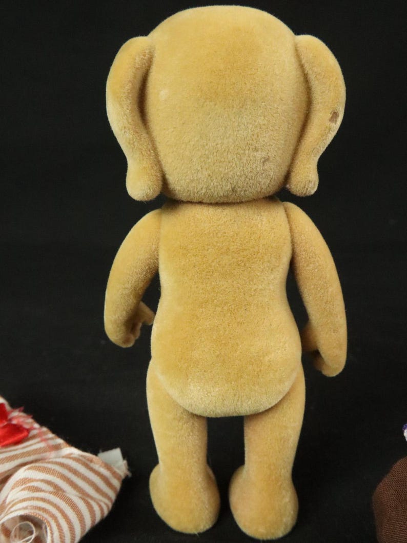 May include: A tan, flocked, anthropomorphic animal figurine standing upright. The figure has rounded ears, a smooth, textured surface, and a simple, upright posture. Beside the figurine lies a small, striped fabric garment with a red bow.