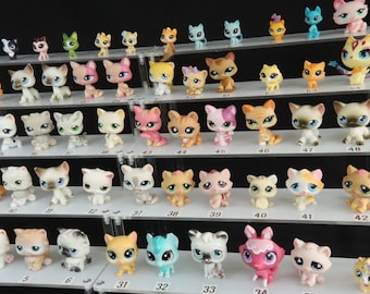 May include: A collection of numerous small, stylized cat figurines arranged on a multi-tiered clear acrylic display stand. The figures feature various colors, patterns, and expressions, with small numbered labels placed on the shelves below each item.