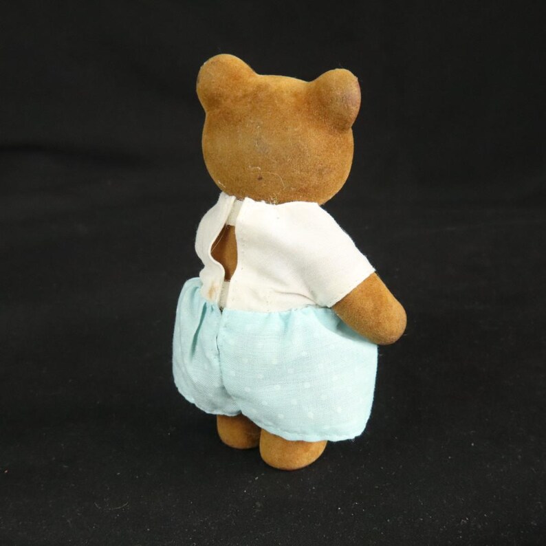 Peut inclure: Une petite figurine d'ours brun floqu&eacute; vue de dos. L'ours porte un haut blanc &agrave; manches courtes ouvert au milieu du dos et un short bleu clair orn&eacute; d'un motif discret &agrave; pois blancs. L'ours poss&egrave;de des oreilles arrondies.
