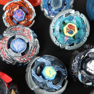May include: A collection of colorful spinning tops in various designs. The tops feature metallic and plastic components, with vibrant colors like red, blue, orange, and silver. Each top has a central emblem and intricate detailing.