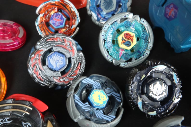 May include: A collection of colorful spinning tops in various designs. The tops feature metallic and plastic components, with vibrant colors like red, blue, orange, and silver. Each top has a central emblem and intricate detailing.