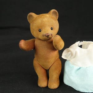Peut inclure: Un ours en peluche floqu&eacute; marron avec des yeux sombres et un petit nez, debout avec les bras tendus. &Agrave; c&ocirc;t&eacute; de l'ours se trouve un petit v&ecirc;tement compos&eacute; d'un haut blanc et d'une jupe bleu clair avec un motif discret &agrave; pois blancs.