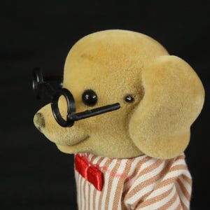 May include: A flocked tan dog figurine wearing black round-rimmed glasses and a white shirt with thin brown vertical stripes, featuring a small red bow tie at the collar. The figure has a smooth, textured surface and black plastic eyes.