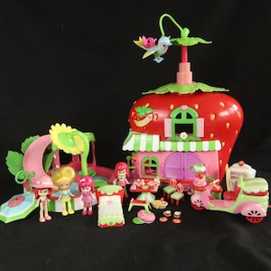 May include: A Strawberry Shortcake playset featuring a large red strawberry-shaped house with green leaves, a pink scooter, a slide, and several small dolls. Accessories include a bed, a table with chairs, a small bird figure, and tiny dishes.