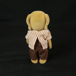 May include: A small, anthropomorphic animal figurine with tan, flocked fur. The figure is wearing a short-sleeved shirt with a white and brown vertical stripe pattern and solid dark brown trousers. The figurine is viewed from the back.