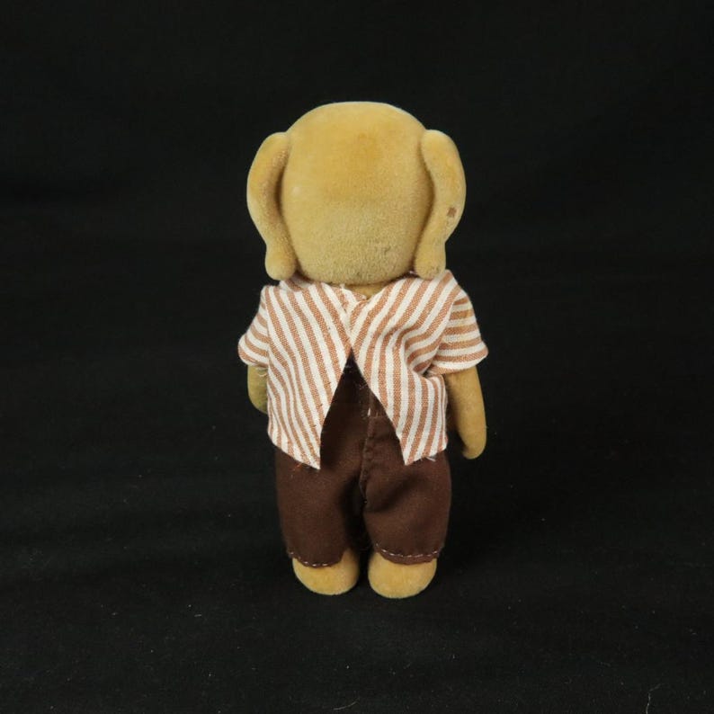 May include: A small, anthropomorphic animal figurine with tan, flocked fur. The figure is wearing a short-sleeved shirt with a white and brown vertical stripe pattern and solid dark brown trousers. The figurine is viewed from the back.