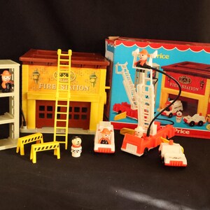 Pode incluir: Um conjunto de brinquedos de estação de bombeiros Fisher-Price vintage. O conjunto inclui um edifício de estação de bombeiros, uma escada, uma torre, carros de bombeiros e figuras de bombeiros. A caixa é visível no fundo. A estação de bombeiros é amarela e castanha, com a palavra "Fire Station".