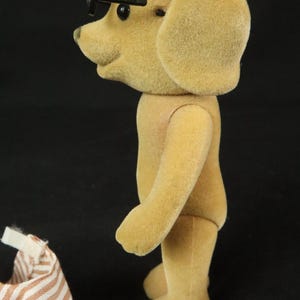 May include: A tan, flocked dog figurine standing in profile, featuring black eyes and small black plastic glasses perched on its snout. Beside the figurine is a piece of white and brown striped fabric clothing with a small white velcro tab.