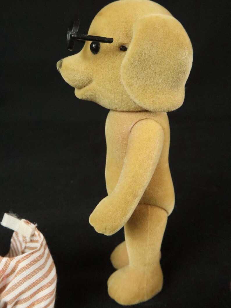 May include: A tan, flocked dog figurine standing in profile, featuring black eyes and small black plastic glasses perched on its snout. Beside the figurine is a piece of white and brown striped fabric clothing with a small white velcro tab.