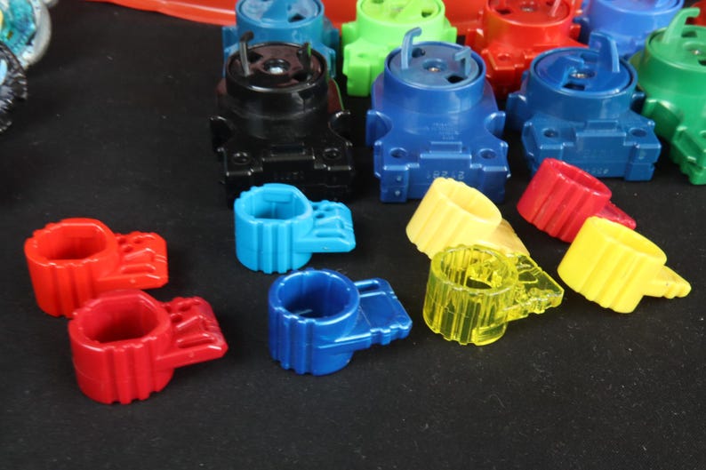 May include: A collection of colorful plastic toy parts in various colors, including red, blue, green, yellow, and black. The parts include cylindrical rings and larger, blocky components with pegs and slots, suggesting a construction or assembly toy.