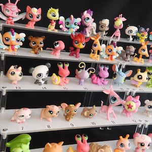 Op de afbeelding: Een verzameling kleine, gestileerde plastic dierenfiguurtjes met grote ogen, gerangschikt op een transparante acryl displaystandaard met meerdere niveaus. Elke plank is voorzien van genummerde labels van 2 tot 20 met diverse kleurrijke wezens.