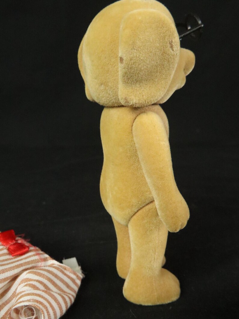 May include: A tan, flocked, anthropomorphic dog figurine standing in profile. The figure has a smooth, fuzzy texture and is wearing black plastic glasses. Beside the figurine lies a piece of fabric with a brown and white striped pattern.