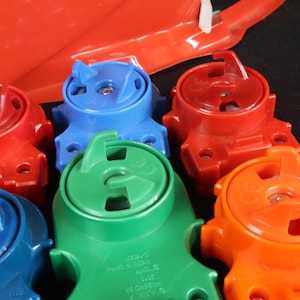 May include: A collection of colorful plastic toy parts in red, blue, green, and orange. The parts have a circular design with a handle and a central mechanism. A red plastic piece is visible in the background.