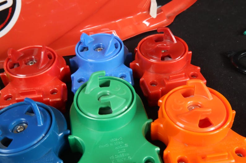 May include: A collection of colorful plastic toy parts in red, blue, green, and orange. The parts have a circular design with a handle and a central mechanism. A red plastic piece is visible in the background.