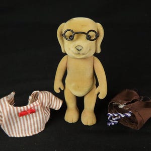 May include: A tan, flocked dog figurine wearing black round-rimmed glasses. Beside the figurine are two pieces of clothing: a white and brown striped shirt with a small red bow and a pair of brown trousers with a blue and white striped drawstring.