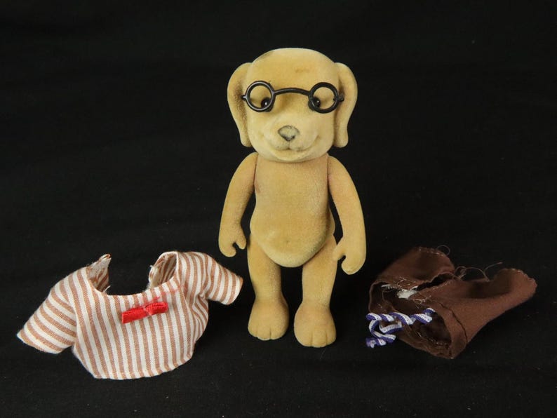 May include: A tan, flocked dog figurine wearing black round-rimmed glasses. Beside the figurine are two pieces of clothing: a white and brown striped shirt with a small red bow and a pair of brown trousers with a blue and white striped drawstring.