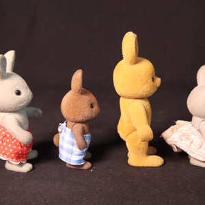 Może przedstawiać: Cztery figurki kr&oacute;liczk&oacute;w zabawek w rzędzie. Kr&oacute;liczki są w r&oacute;żnych kolorach, w tym szarym, brązowym i ż&oacute;łtym. Dwa kr&oacute;liki mają na sobie sukienki, jedna czerwoną w białe grochy, a druga r&oacute;żową we wzory kwiatowe. Kolejny ma niebiesko-białe ogrodniczki.