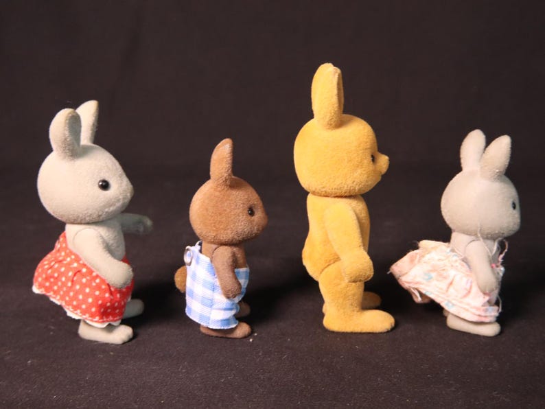 Może przedstawiać: Cztery figurki kr&oacute;liczk&oacute;w zabawek w rzędzie. Kr&oacute;liczki są w r&oacute;żnych kolorach, w tym szarym, brązowym i ż&oacute;łtym. Dwa kr&oacute;liki mają na sobie sukienki, jedna czerwoną w białe grochy, a druga r&oacute;żową we wzory kwiatowe. Kolejny ma niebiesko-białe ogrodniczki.