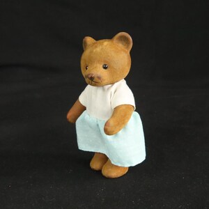 Peut inclure: Une petite figurine d'ours brun floqu&eacute; debout. L'ours a des yeux noirs et un nez brun fonc&eacute;. Il porte un haut blanc &agrave; manches courtes associ&eacute; &agrave; une jupe bleu clair orn&eacute;e d'un motif subtil de pois blancs r&eacute;p&eacute;t&eacute;s sur tout le tissu.