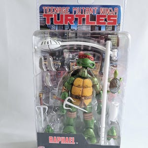 Puede incluir: Figura de acción de Raphael de las Tortugas Ninja, empaquetada en una funda de plástico transparente. La figura es verde con una máscara roja y un caparazón amarillo. Los accesorios incluyen un sai, un bastón y otras armas. El embalaje presenta las palabras "Teenage Mutant Ninja Turtles" y "Raphael".