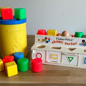 Op de afbeelding: Een vintage Fisher-Price vormsorteerspeelgoed. De set bevat een gele cilinder met een blauwe rand en een witte vormsorteerder met diverse gekleurde blokken. De vormsorteerder heeft een verscheidenheid aan vormen, waaronder een cirkel, een vierkant, een rechthoek en een driehoek.