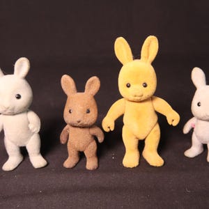 Może przedstawiać: Cztery małe, flokowane figurki kr&oacute;lik&oacute;w w kolorach białym, brązowym i ż&oacute;łtym. Każdy ma duże uszy, małe czarne oczy i pozycję stojącą. To zabawki lub przedmioty kolekcjonerskie.