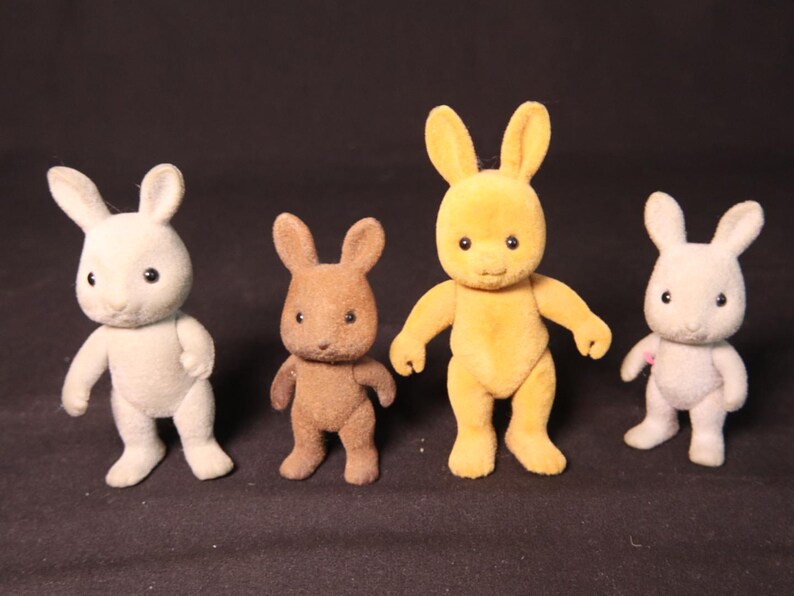 Może przedstawiać: Cztery małe, flokowane figurki kr&oacute;lik&oacute;w w kolorach białym, brązowym i ż&oacute;łtym. Każdy ma duże uszy, małe czarne oczy i pozycję stojącą. To zabawki lub przedmioty kolekcjonerskie.