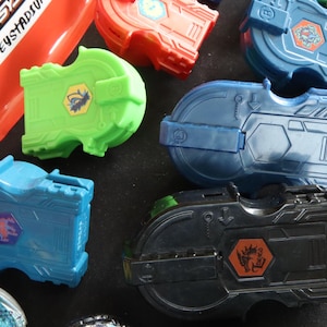 May include: A collection of colorful toy launchers in various shades of blue, green, red, and black. Each launcher features a unique design with hexagonal emblems and intricate details. The image also includes red and yellow rings.