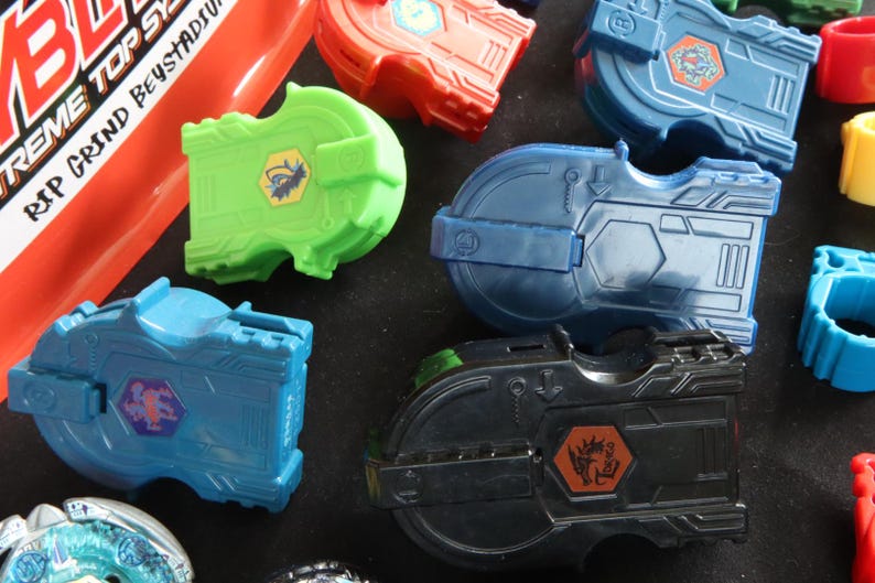 May include: A collection of colorful toy launchers in various shades of blue, green, red, and black. Each launcher features a unique design with hexagonal emblems and intricate details. The image also includes red and yellow rings.