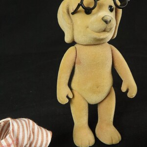 May include: A tan, flocked dog figurine with a smooth, velvety texture, standing upright with articulated limbs. The dog has black, round eyes, a small black nose, and is wearing a pair of black, circular-rimmed glasses perched on its forehead.