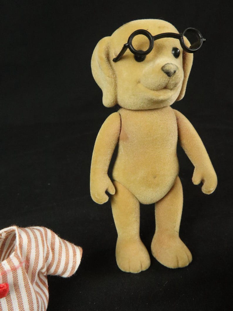 May include: A tan, flocked dog figurine with a smooth, velvety texture, standing upright with articulated limbs. The dog has black, round eyes, a small black nose, and is wearing a pair of black, circular-rimmed glasses perched on its forehead.