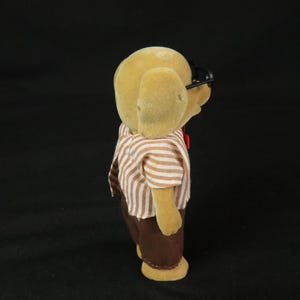 May include: A tan flocked dog figurine wearing black plastic glasses, a white and brown striped short-sleeved shirt, and solid brown trousers. The small anthropomorphic animal figure is viewed from the side, showing its textured surface and clothing.