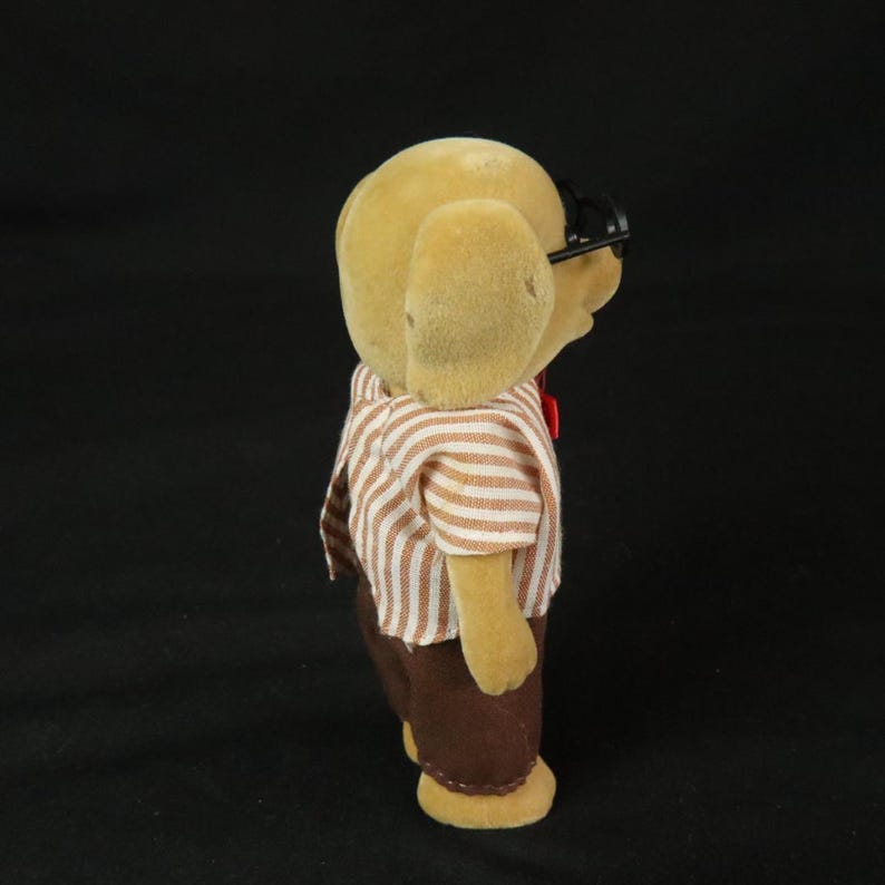 May include: A tan flocked dog figurine wearing black plastic glasses, a white and brown striped short-sleeved shirt, and solid brown trousers. The small anthropomorphic animal figure is viewed from the side, showing its textured surface and clothing.